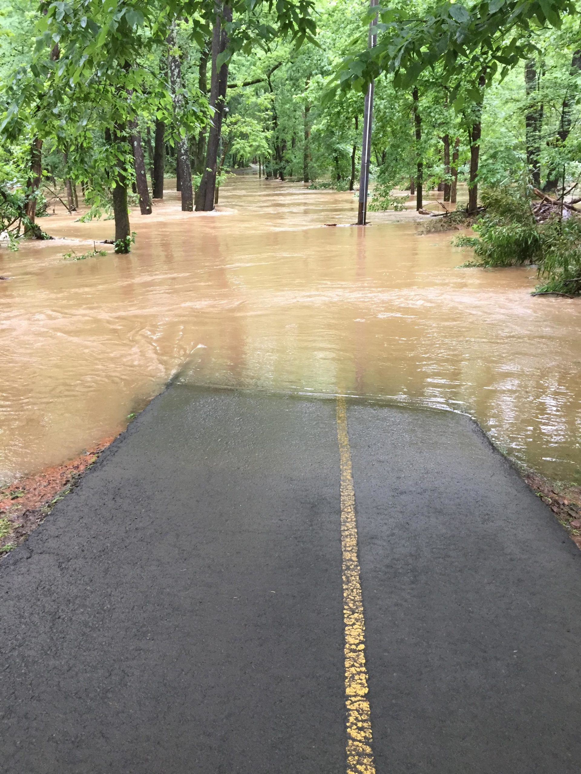 A paved pathway, partially submerged in muddy water, surrounded by lush green trees after heavy rainfall. The scene shows flooded areas on both sides of the path, with visible water level rising above the ground. Spadra Creek Nature Trail mountain bike trail.
