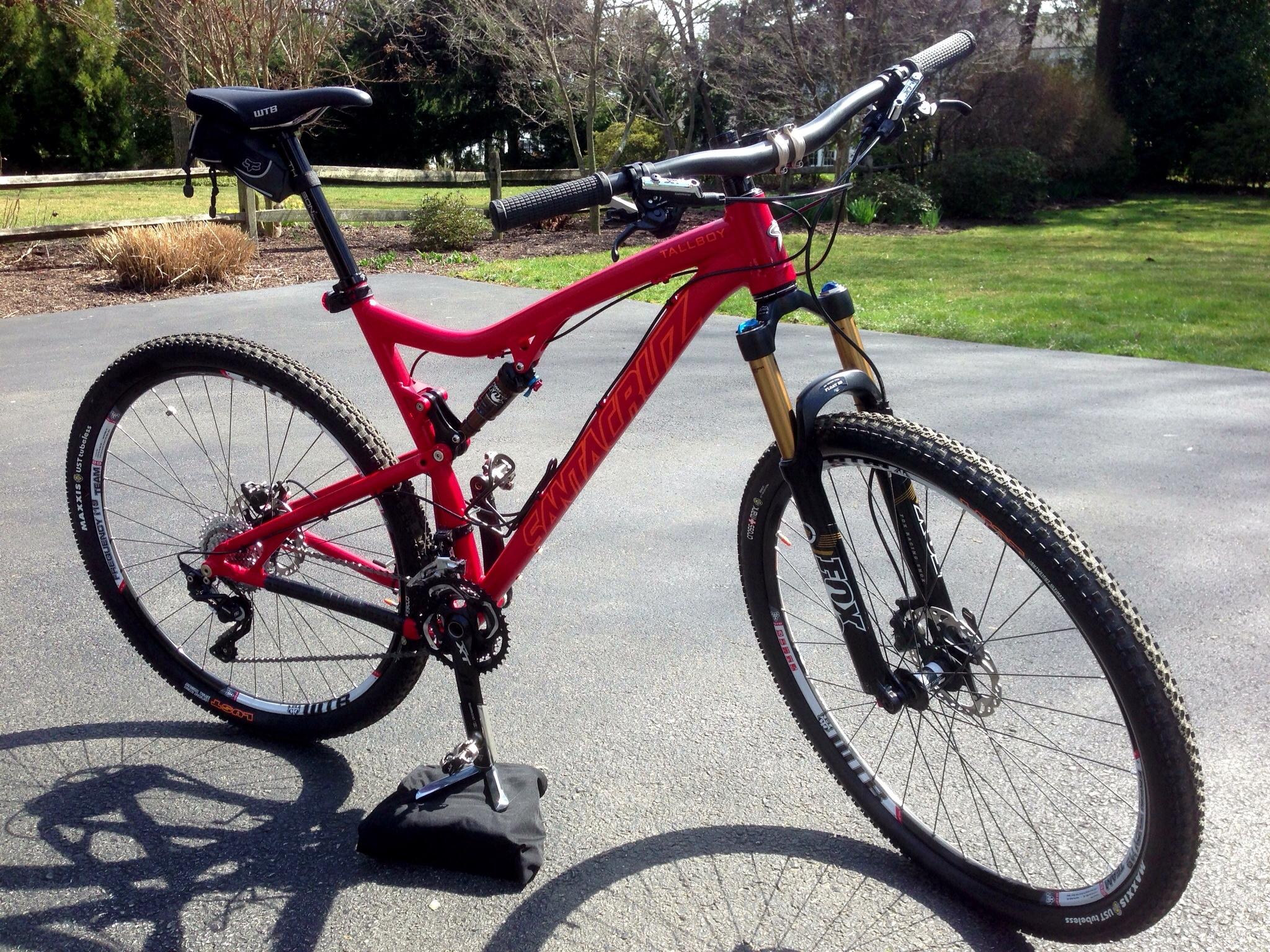 Santa Cruz Tallboy: A vibrant red mountain bike with a sleek design parked on a paved surface. The bike features a front suspension fork, disc brakes, and a gear system. A grassy area and trees are visible in the background, suggesting an outdoor setting.