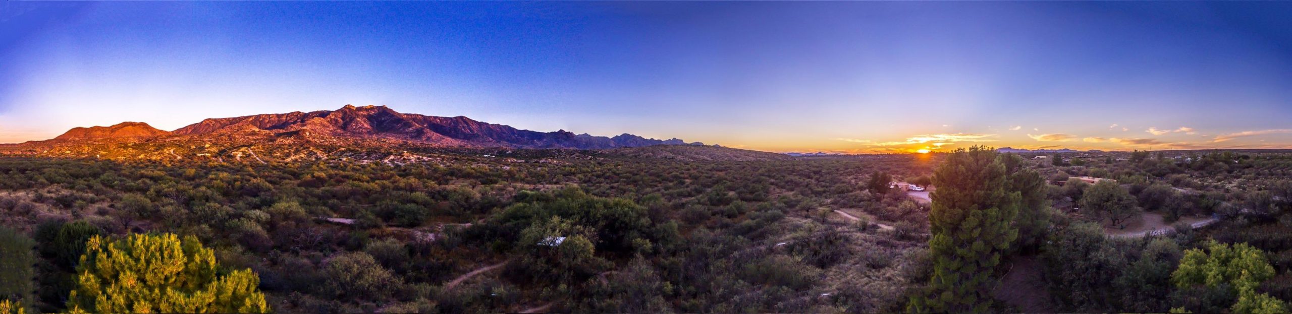 A panoramic view of a desert landscape during sunset, featuring rugged mountains in the background, a colorful sky transitioning from orange to deep blue, and sparse vegetation in the foreground. The scene captures the serene beauty of a natural desert environment. 50-year Trail / Golder Ranch mountain bike trail.