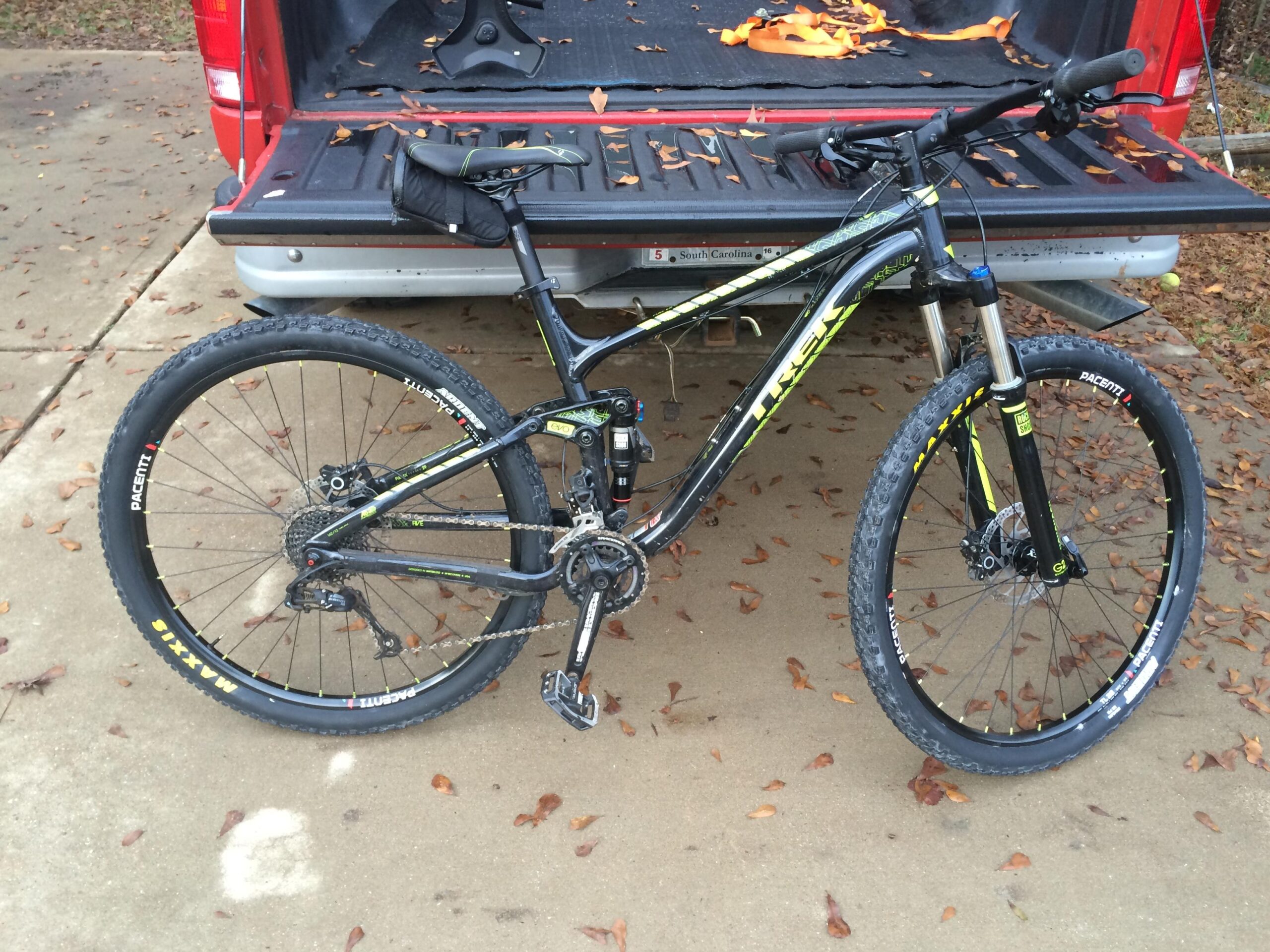 Trek Fuel EX 5 29: A black and green mountain bike resting on a concrete surface next to the open tailgate of a red pickup truck. The bike features rugged tires and a front suspension fork, with fallen leaves scattered around.