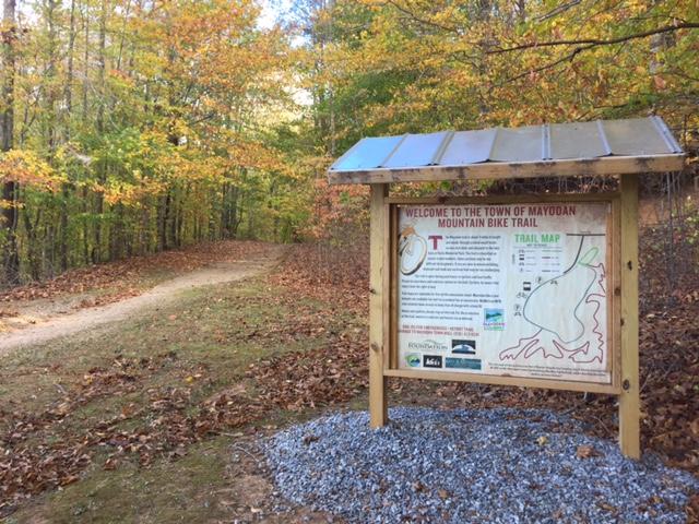 Sign welcoming visitors to the Mayodan Mountain Bike Trail, featuring a trail map and information about the area's biking routes, surrounded by colorful autumn trees and a dirt path leading into the woods. Farris Park mountain bike trail.