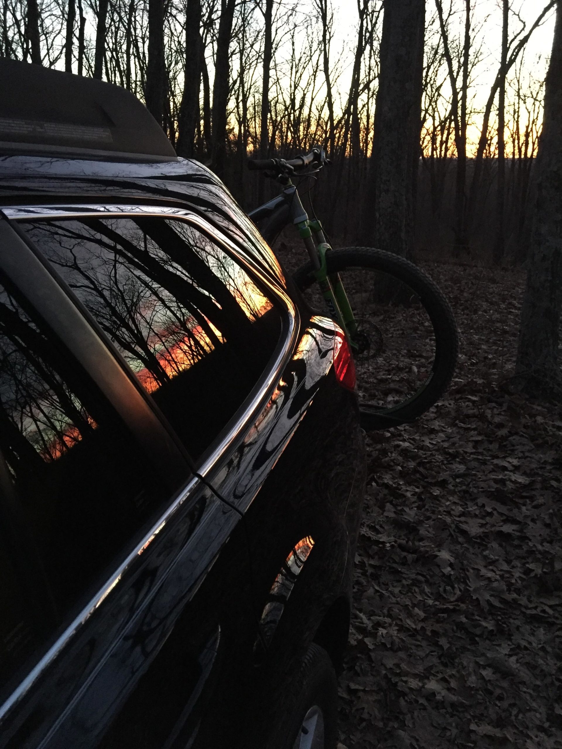 A black vehicle parked in a wooded area at dusk, reflecting a colorful sunset in its windows. A mountain bike is mounted on the back of the vehicle, surrounded by trees with bare branches. Leaves cover the ground, creating a natural setting. Beulah mountain bike trail.