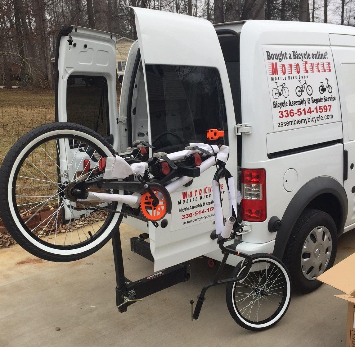 A white van with the logo of a mobile bike shop, "Moto Cycle," parked outdoors. The back of the van is open, displaying a bike partially assembled with one wheel attached, hanging on a bike rack. Text on the van promotes bicycle assembly and repair services. A grassy area is visible in the background.