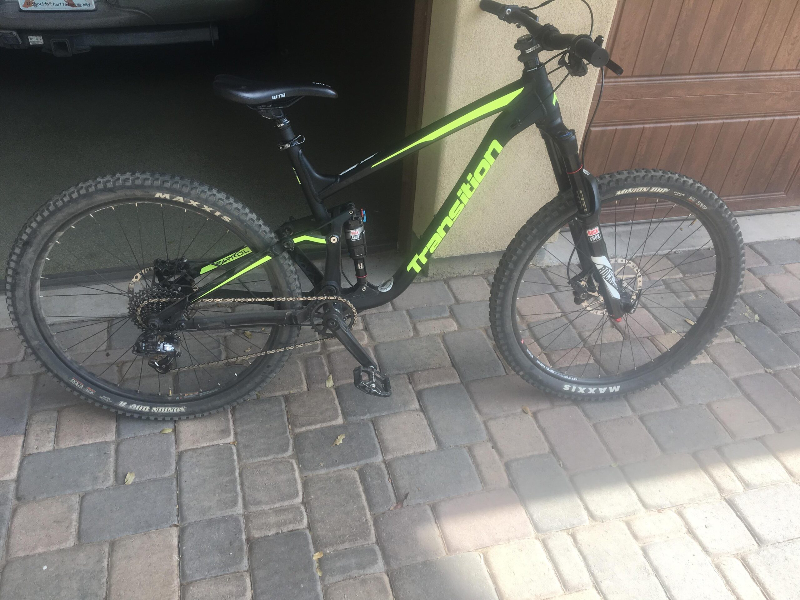 Transition Patrol: A black and green mountain bike parked on a stone driveway, partially visible in front of a garage. The bike features knobby tires, a suspension fork, and a modern frame design, with visible branding on the frame.