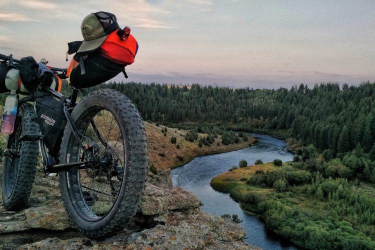 A mountain bike with large tires and packed gear rests on a rocky ledge, overlooking a winding river surrounded by dense green forests. The sky is softly lit with pastel colors, suggesting either dawn or dusk.