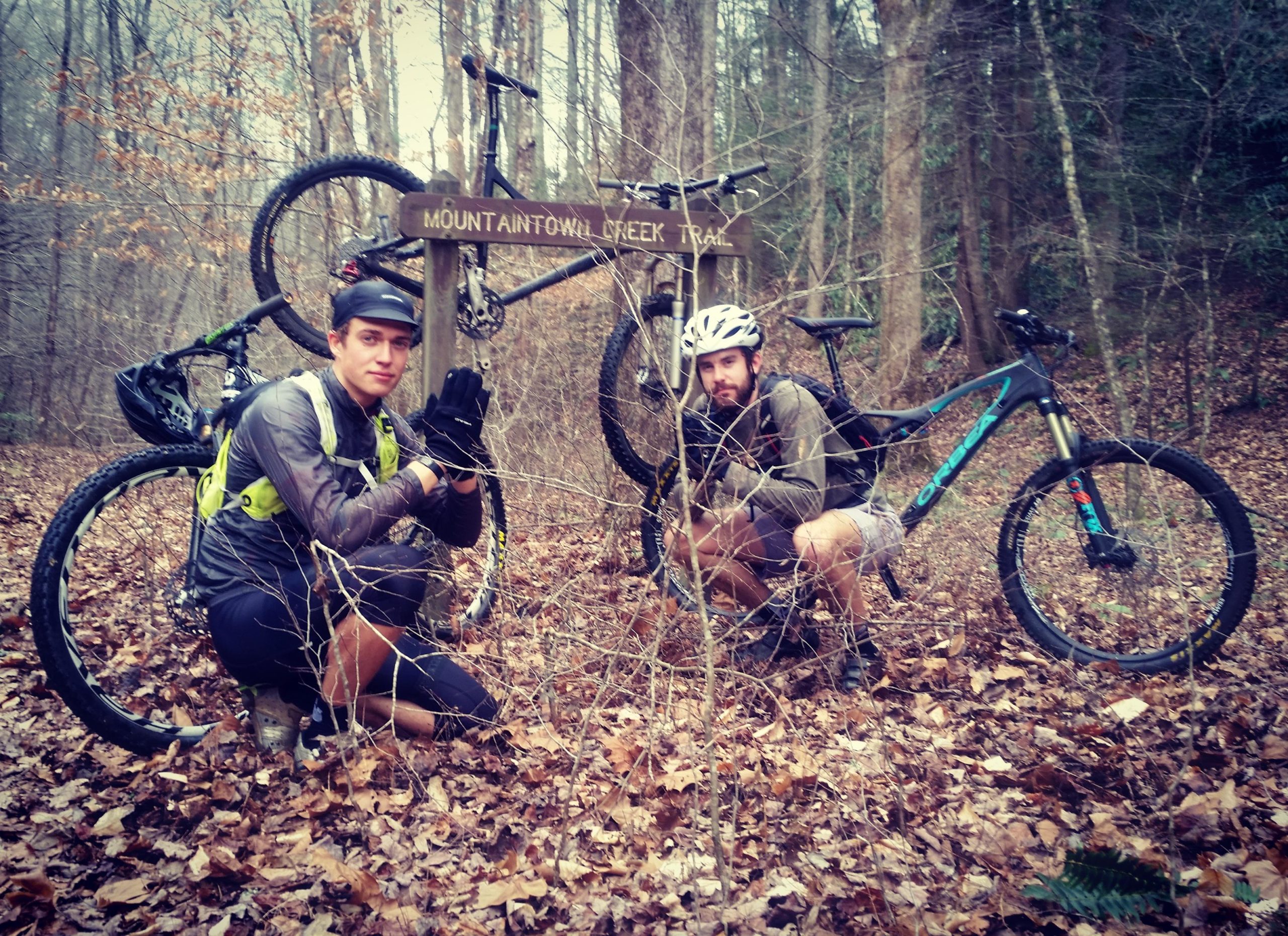 Two mountain bikers crouch next to a trail sign labeled "Mountaintown Creek Trail," surrounded by wooded terrain and fallen leaves. One biker wears a helmet and gloves with a hydration pack, while the other has a beard and a casual outfit. Their bikes are leaning against the sign behind them. Pinhoti Trail: Mountaintown Creek Segment mountain bike trail.