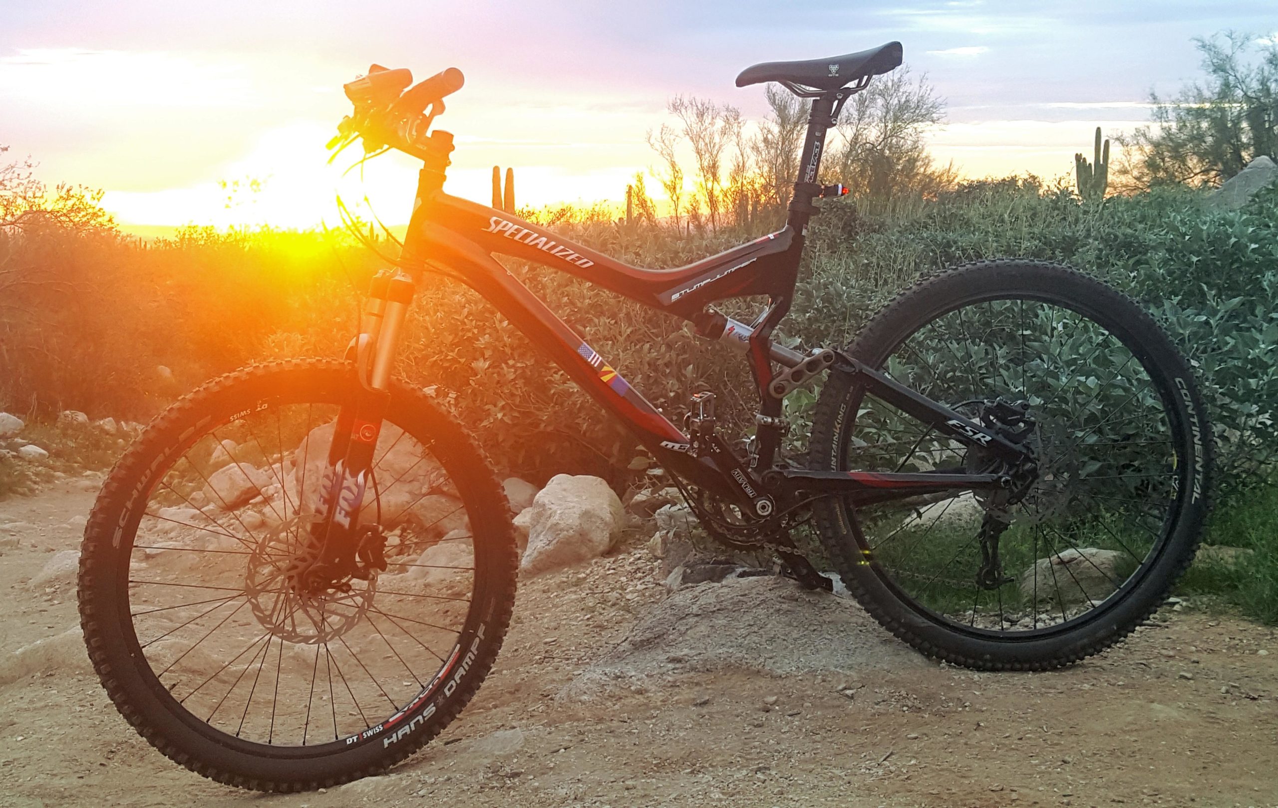 A black mountain bike positioned on a dirt path with vibrant sunset light in the background, featuring desert vegetation and rocks. The scene captures the essence of outdoor adventure and cycling in nature. White Tanks Regional Park Trails mountain bike trail.