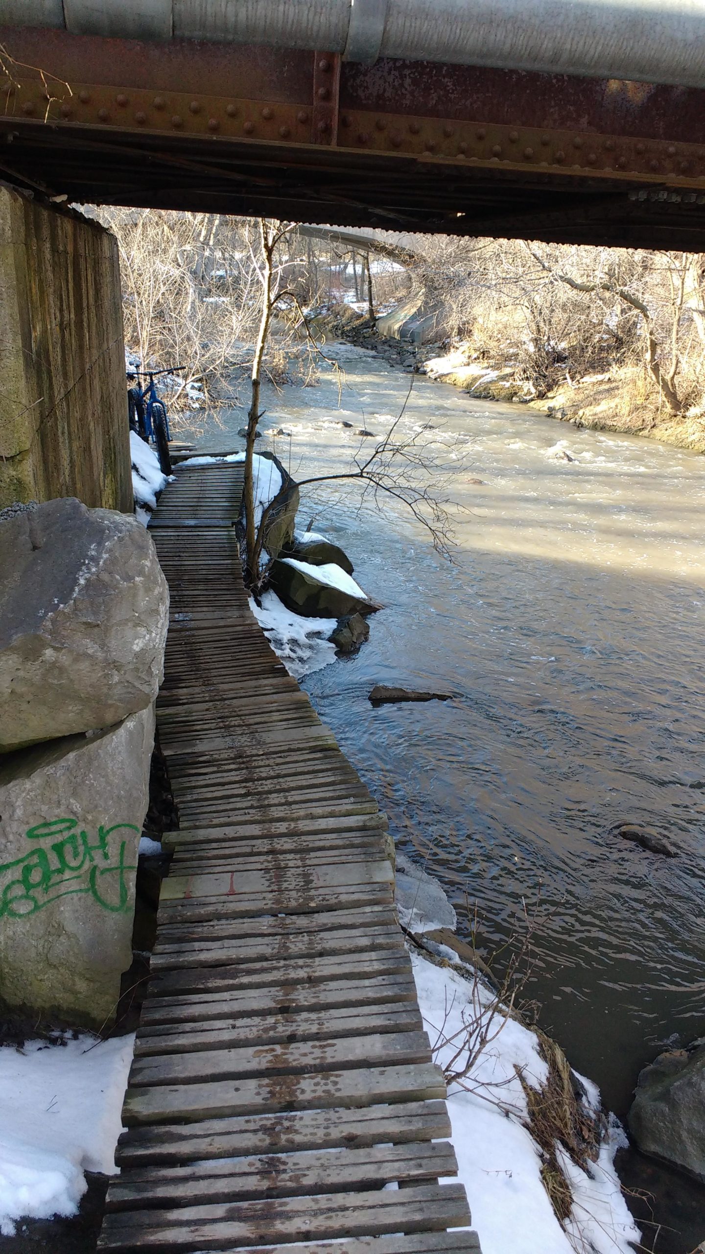 A wooden walkway runs alongside a river, with snow covering some areas along the banks. A rocky ledge and a concrete structure are visible, alongside a rusted metal bridge overhead. The river flows gently, reflecting the winter landscape. Don Valley mountain bike trail.