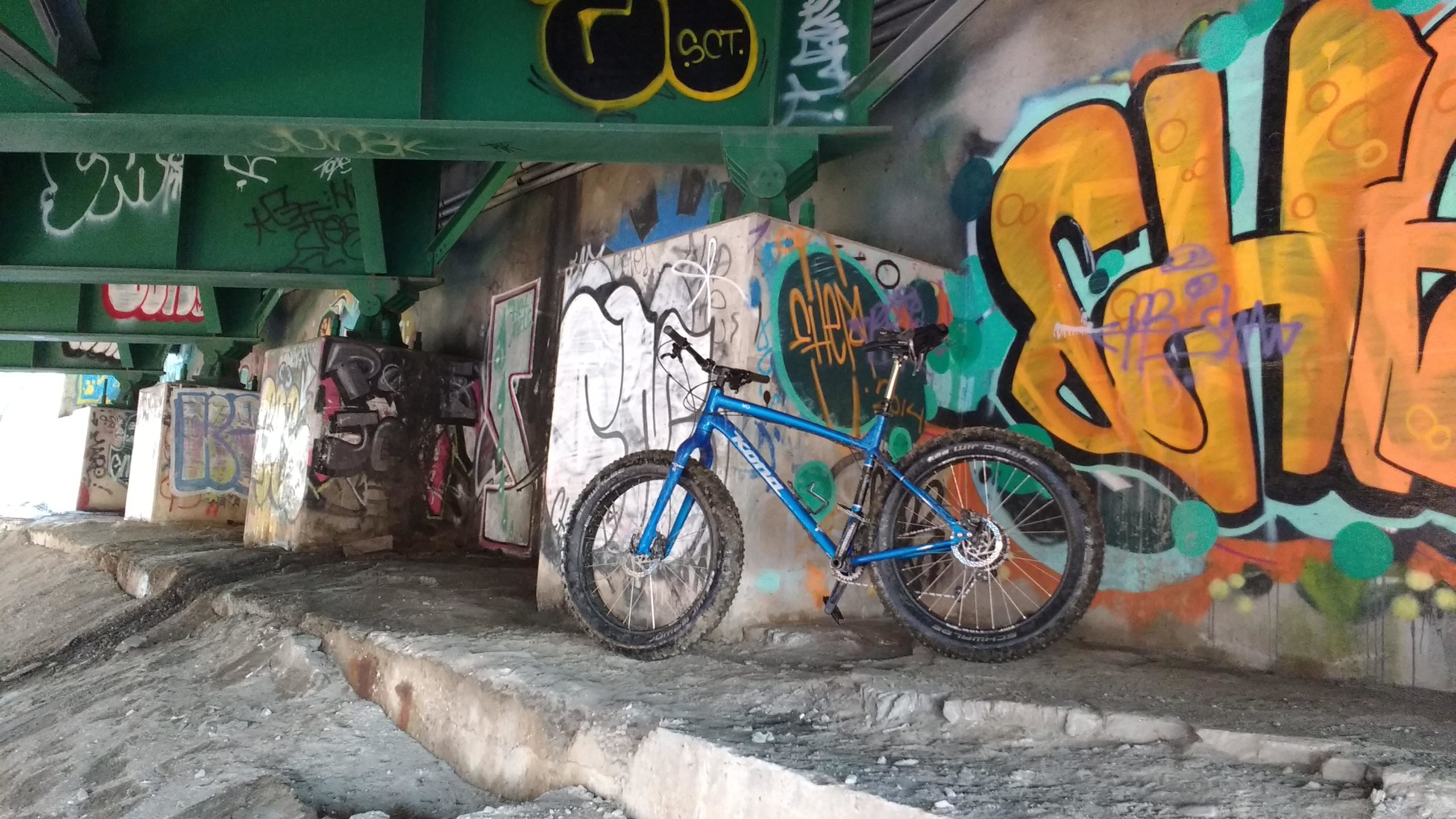 A blue mountain bike is leaning against a graffiti-covered wall beneath a green bridge. The ground is uneven and rocky, with various vibrant graffiti designs in the background. Don Valley mountain bike trail.