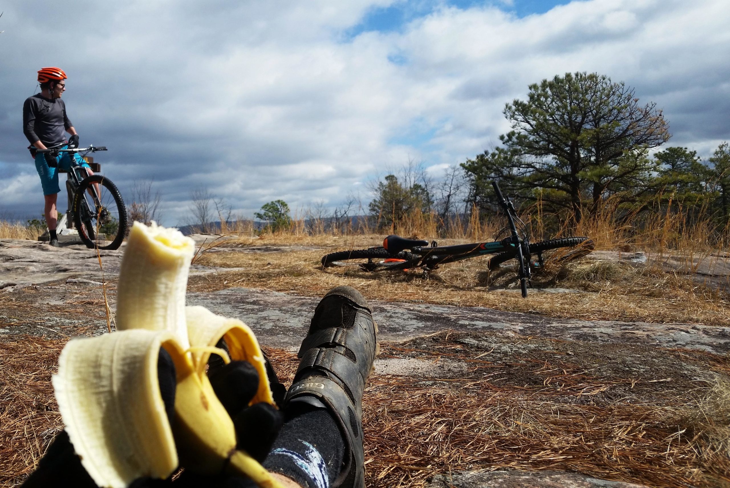 A mountain biker stands in the background near a rocky outcrop, looking out over a scenic view. In the foreground, a hand holds a partially eaten banana, with a bike resting on the ground nearby. The landscape features grass and sparse trees under a cloudy sky. DuPont State Recreational Forest mountain bike trail.
