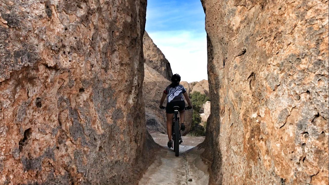 A mountain biker navigating through a narrow rock formation, framed by towering stone walls, with a clear blue sky in the background. City Of Rocks mountain bike trail.