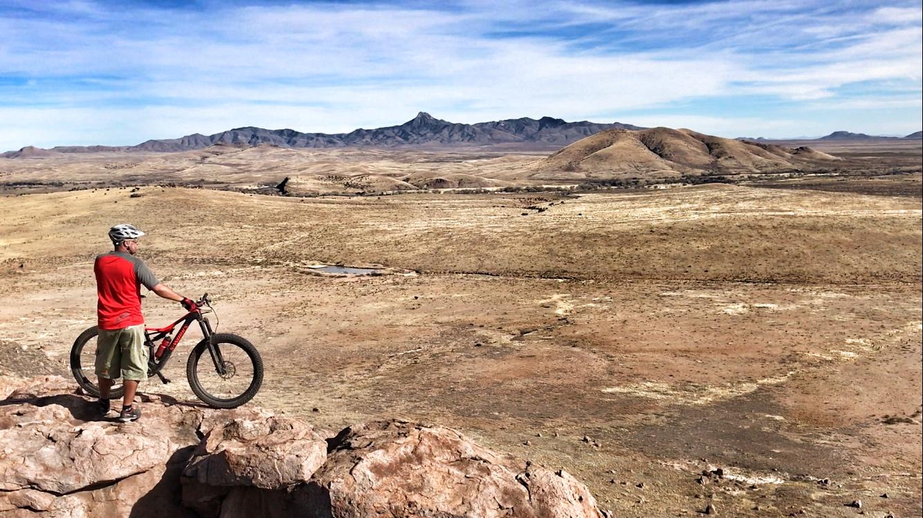 A person in a red sleeveless shirt and a helmet stands beside a mountain bike on a rocky outcrop, overlooking a vast, arid landscape with rolling hills and distant mountains under a blue sky. City Of Rocks mountain bike trail.