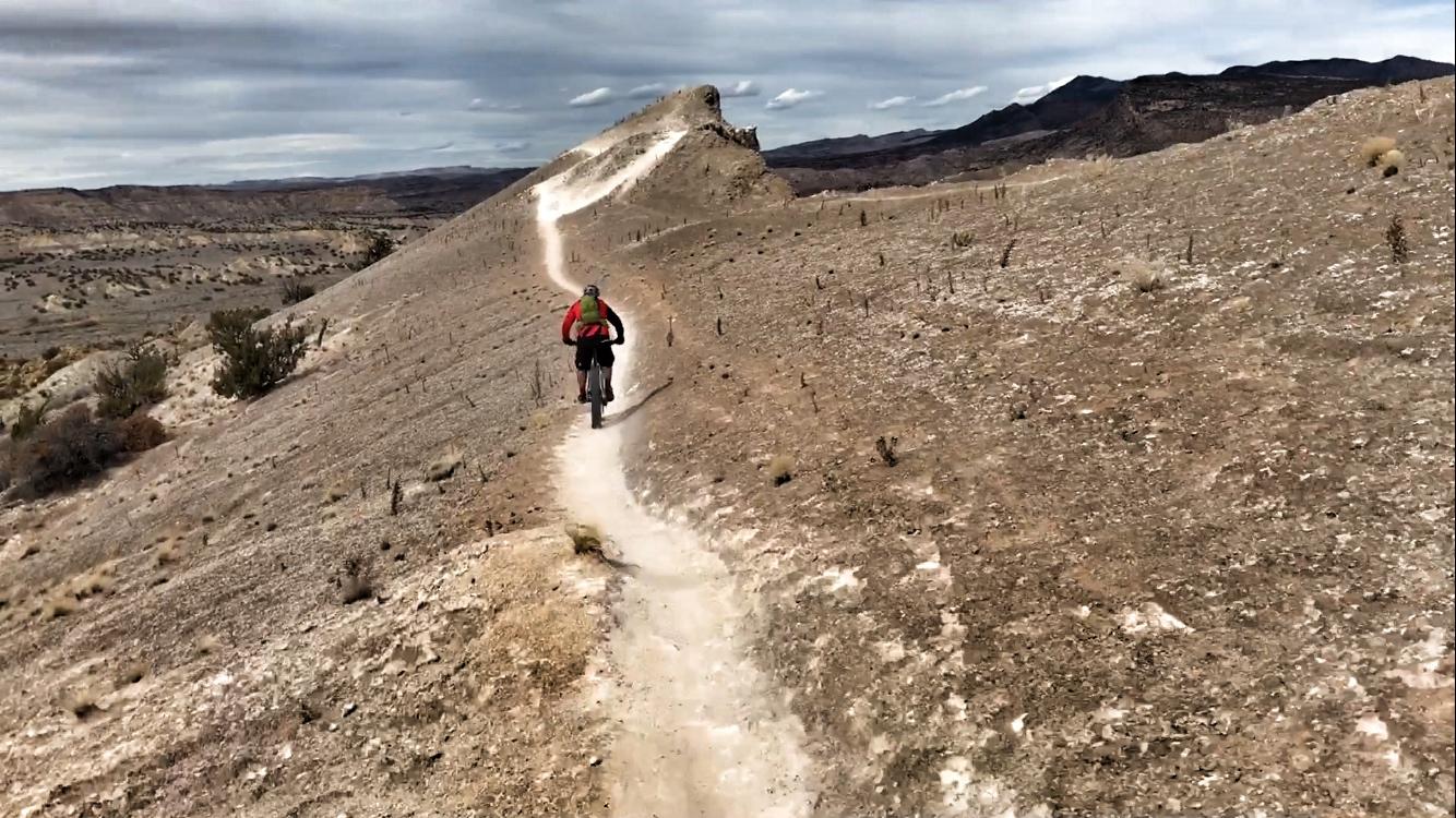 A cyclist rides along a winding dirt trail on a rocky, arid hillside with sparse vegetation. The landscape features distant mountains under a cloudy sky, creating a scenic backdrop for outdoor adventure. White Ridge Bike Trails mountain bike trail.