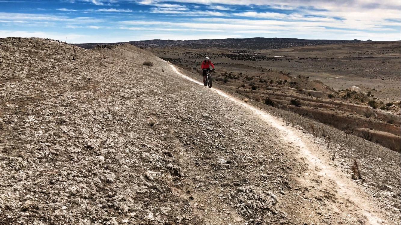 A mountain biker riding along a rocky dirt trail on a hillside, with expansive plains and distant hills in the background under a partly cloudy sky. White Ridge Bike Trails mountain bike trail.