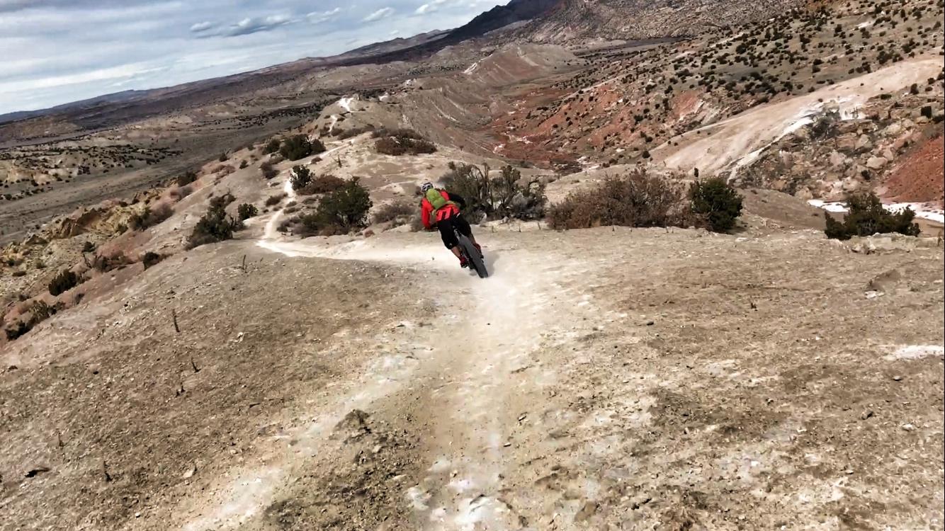 A person riding a mountain bike down a dirt trail on a rocky landscape, surrounded by rolling hills and sparse vegetation under a cloudy sky. White Ridge Bike Trails mountain bike trail.