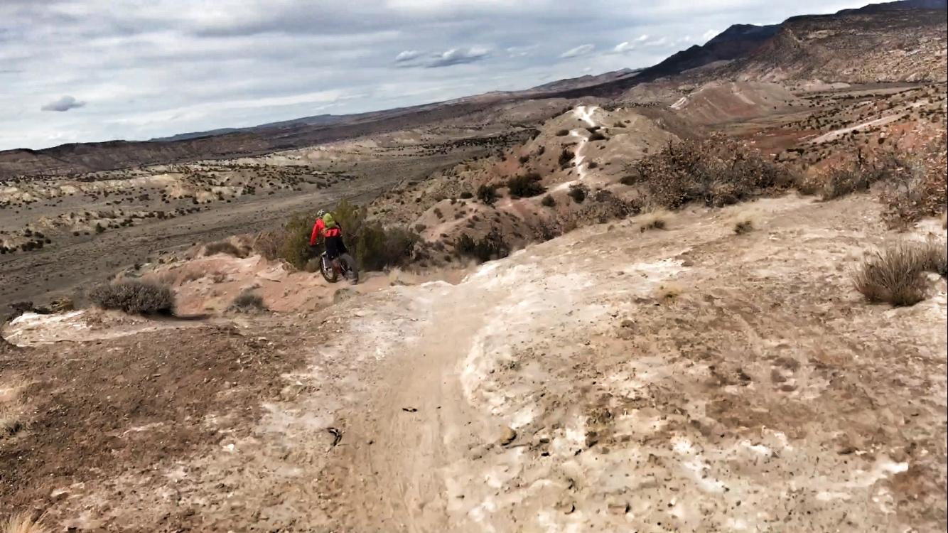 A mountain biker in a red jacket descends a sandy, rocky trail surrounded by a wide-open landscape of hills and sparse vegetation under a cloudy sky. White Ridge Bike Trails mountain bike trail.