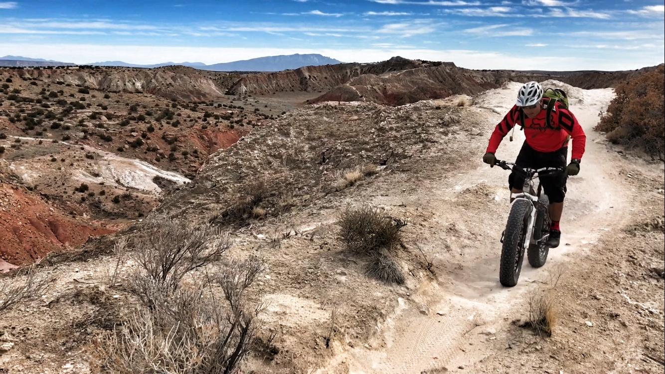 A mountain biker in a red jersey navigates a dirt trail along a rugged landscape, with rocky hills and sparse vegetation in the background under a blue sky. White Ridge Bike Trails mountain bike trail.