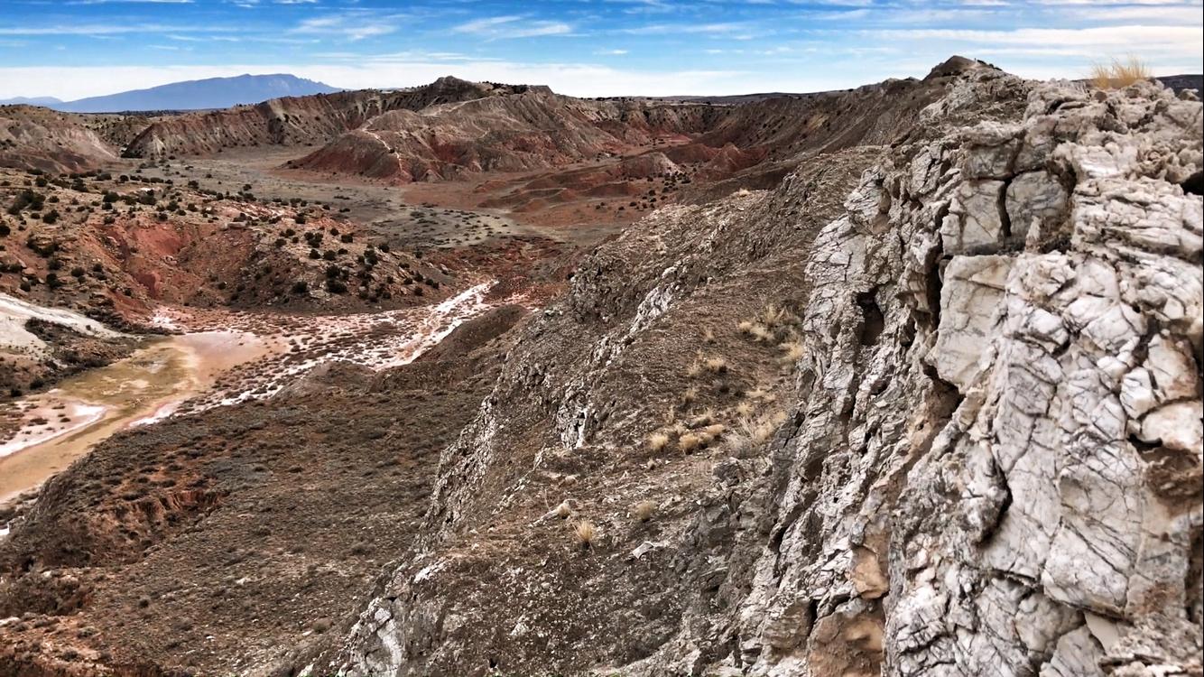 A panoramic view of a rugged, dry landscape featuring layered rock formations and a winding path, with reddish and gray hues. Sparse vegetation dots the scenery under a partly cloudy sky, with distant mountains visible on the horizon. White Ridge Bike Trails mountain bike trail.