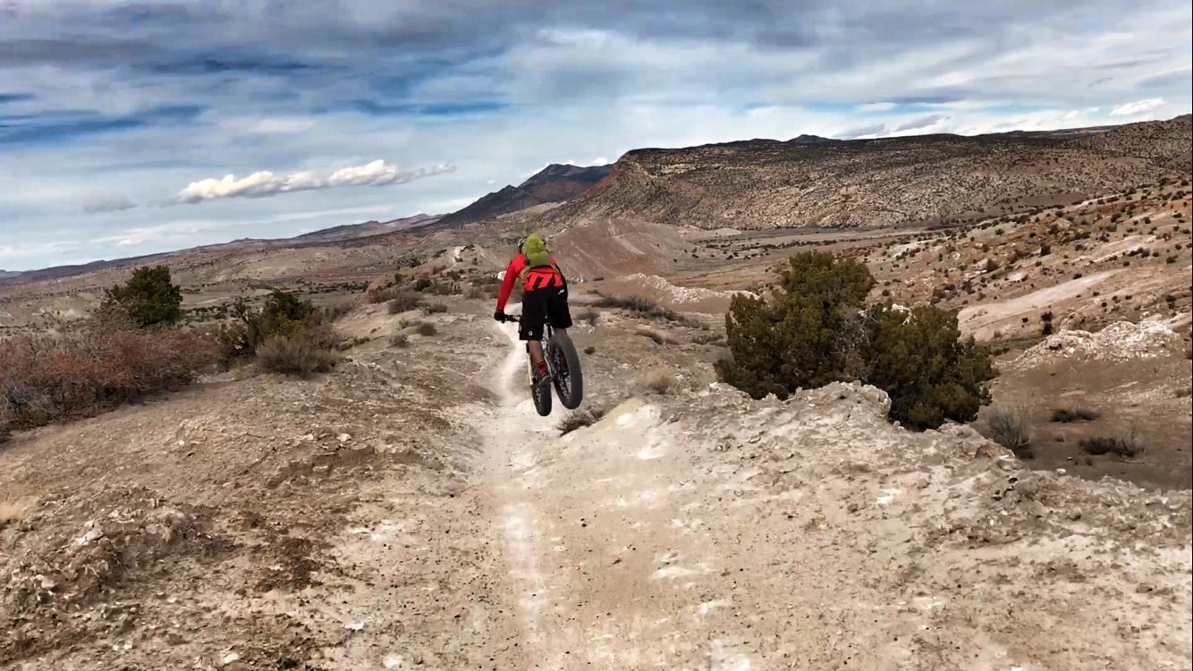 A cyclist dressed in a red jacket and black shorts rides a fat bike along a narrow dirt trail, surrounded by rocky terrain and sparse vegetation. The landscape features rolling hills and mountainous backgrounds under a partly cloudy sky. White Ridge Bike Trails mountain bike trail.