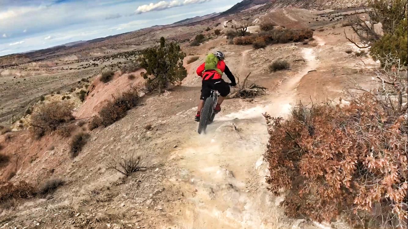 A mountain biker riding down a rocky trail in a desert landscape, with sparse vegetation and hills in the background under a partly cloudy sky. Dust is kicked up from the tires as the rider navigates the terrain. White Ridge Bike Trails mountain bike trail.