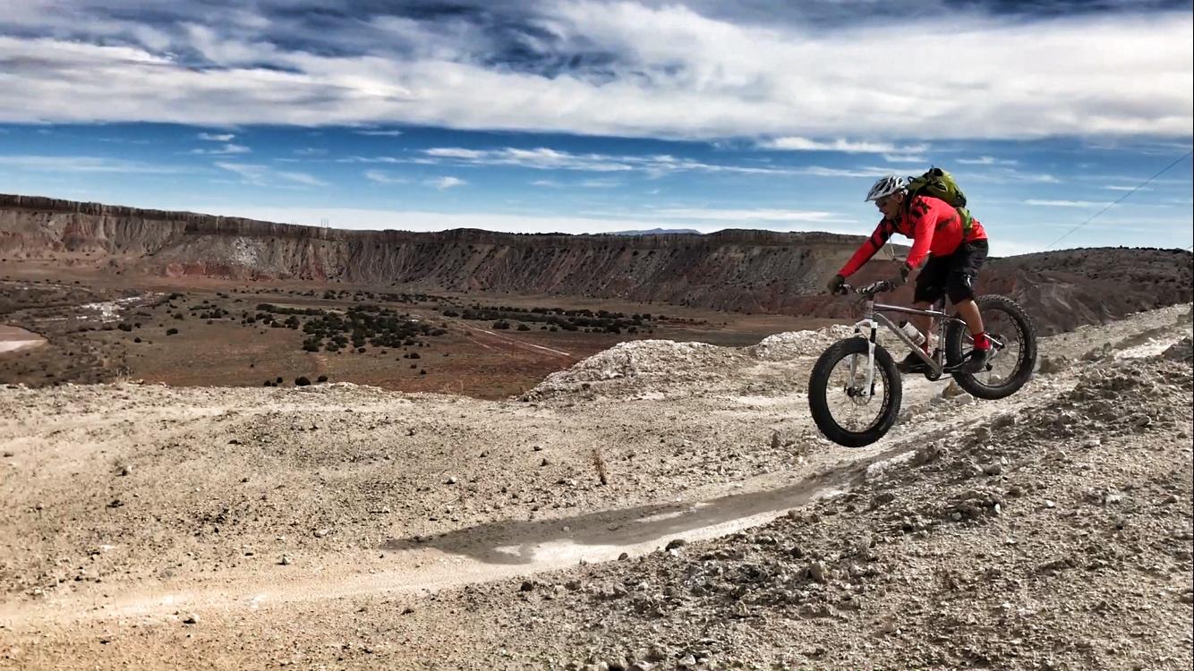 A mountain biker in a red shirt and helmet is mid-jump on a rocky, sandy trail, with a vast canyon landscape in the background under a partly cloudy sky. White Ridge Bike Trails mountain bike trail.