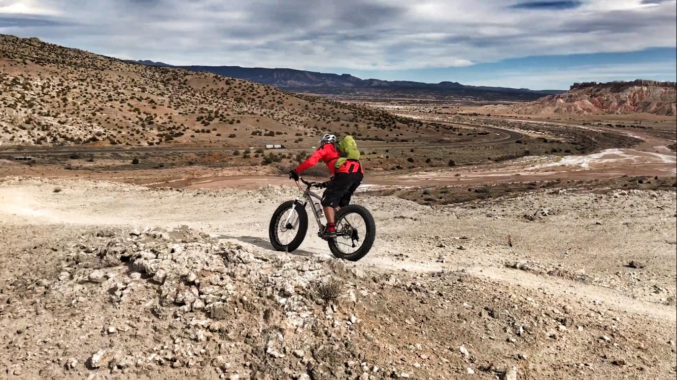 A person riding a fat bike on a rocky trail in a mountainous desert landscape, with hills and winding roads in the background under a partly cloudy sky. White Ridge Bike Trails mountain bike trail.