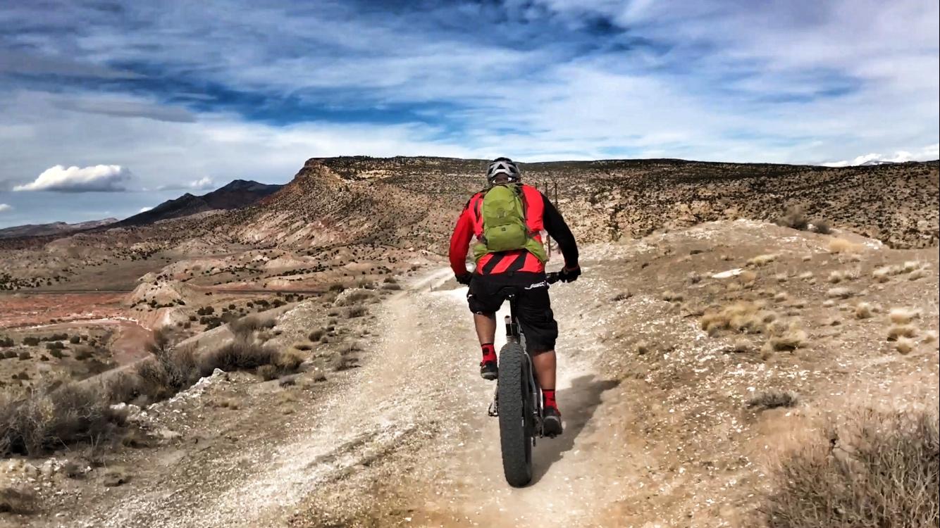 A person riding a fat bike on a dirt trail surrounded by rugged terrain and mountains under a partly cloudy sky. White Ridge Bike Trails mountain bike trail.