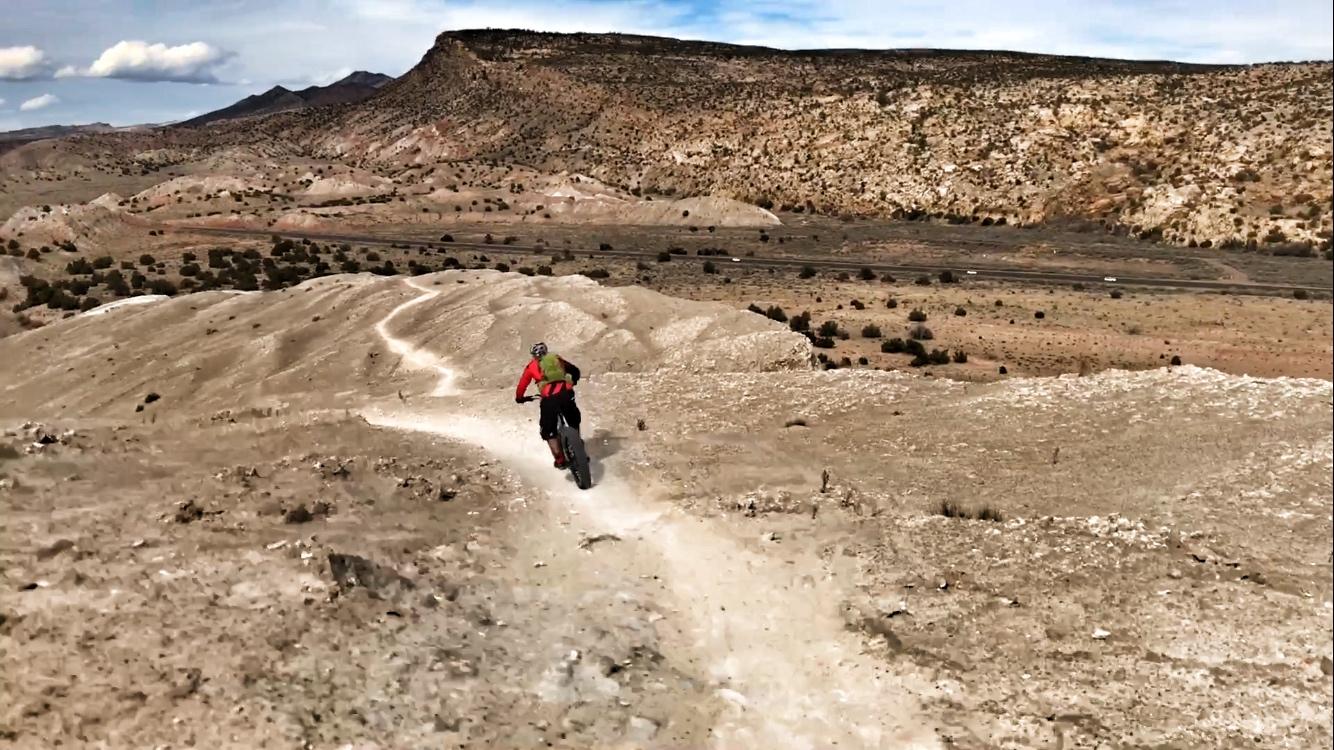 A person riding a mountain bike on a rocky trail in a barren landscape, with hills and sparse vegetation in the background under a partly cloudy sky. White Ridge Bike Trails mountain bike trail.