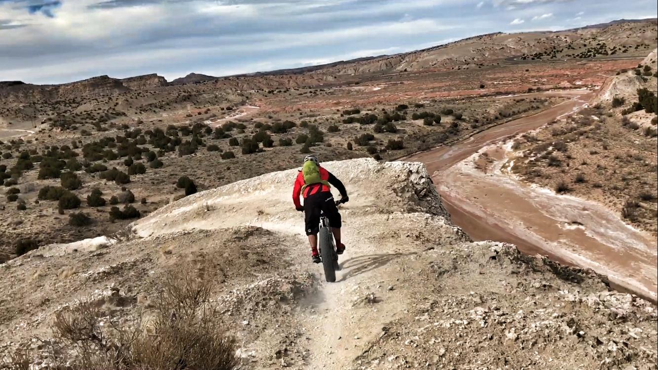 A cyclist riding a fat bike down a dirt trail on a rocky slope, with a panoramic view of a rugged, arid landscape in the background. The scene features hills, sparse vegetation, and a winding path in the valley below, under a cloudy sky. White Ridge Bike Trails mountain bike trail.