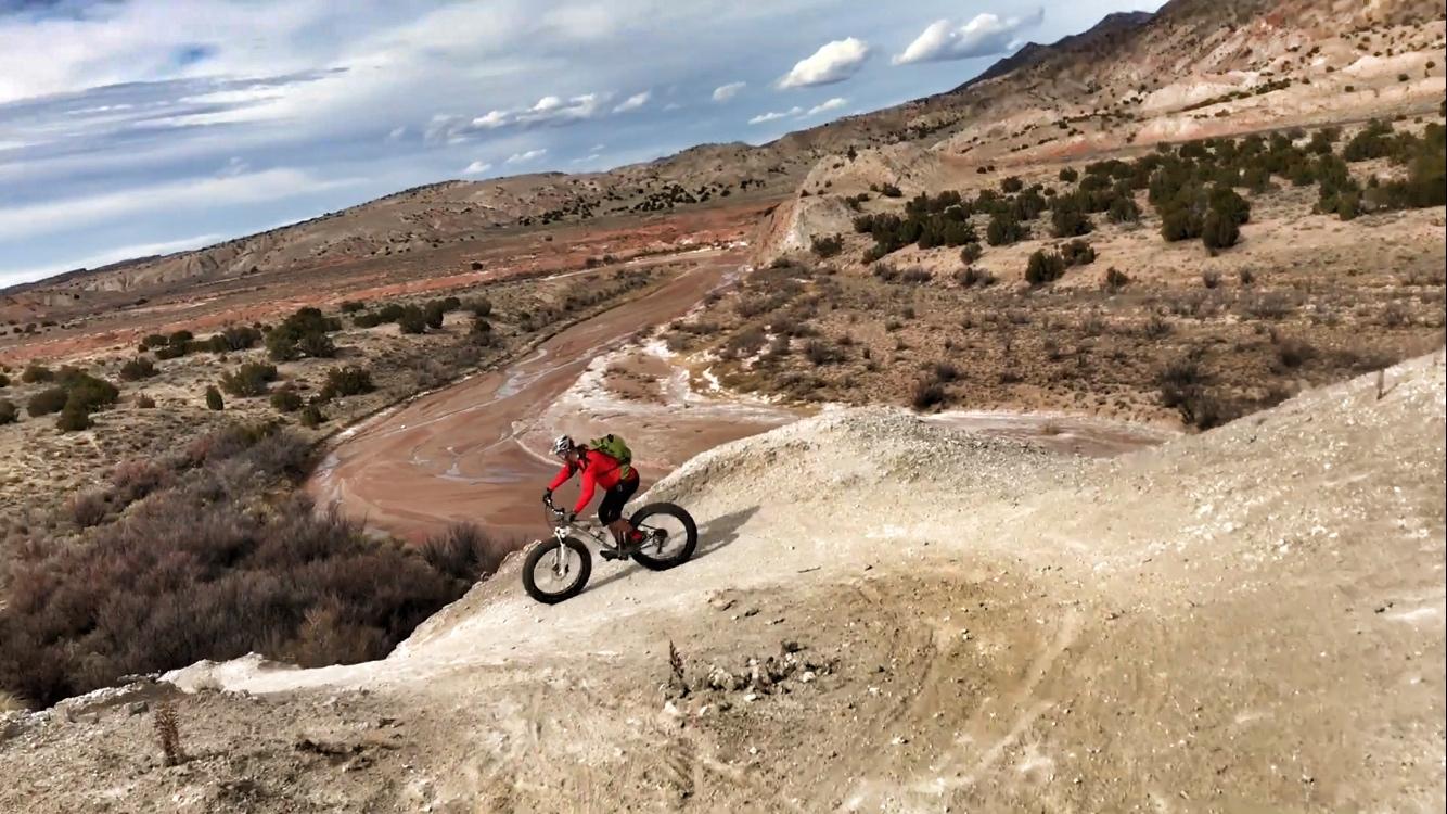A mountain biker in a red jacket rides along a rugged dirt trail in a barren landscape, surrounded by hills and sparse vegetation under a cloudy sky. White Ridge Bike Trails mountain bike trail.