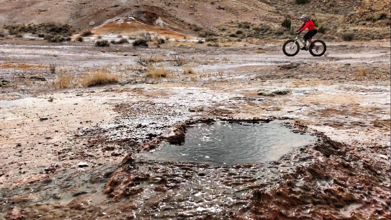 A mountain biker in a red jacket rides on a rocky, barren landscape with patches of water and mineral deposits. In the foreground, a small, reflective pool of water is surrounded by earthy, textured terrain. The background features rolling hills and dry vegetation. White Ridge Bike Trails mountain bike trail.