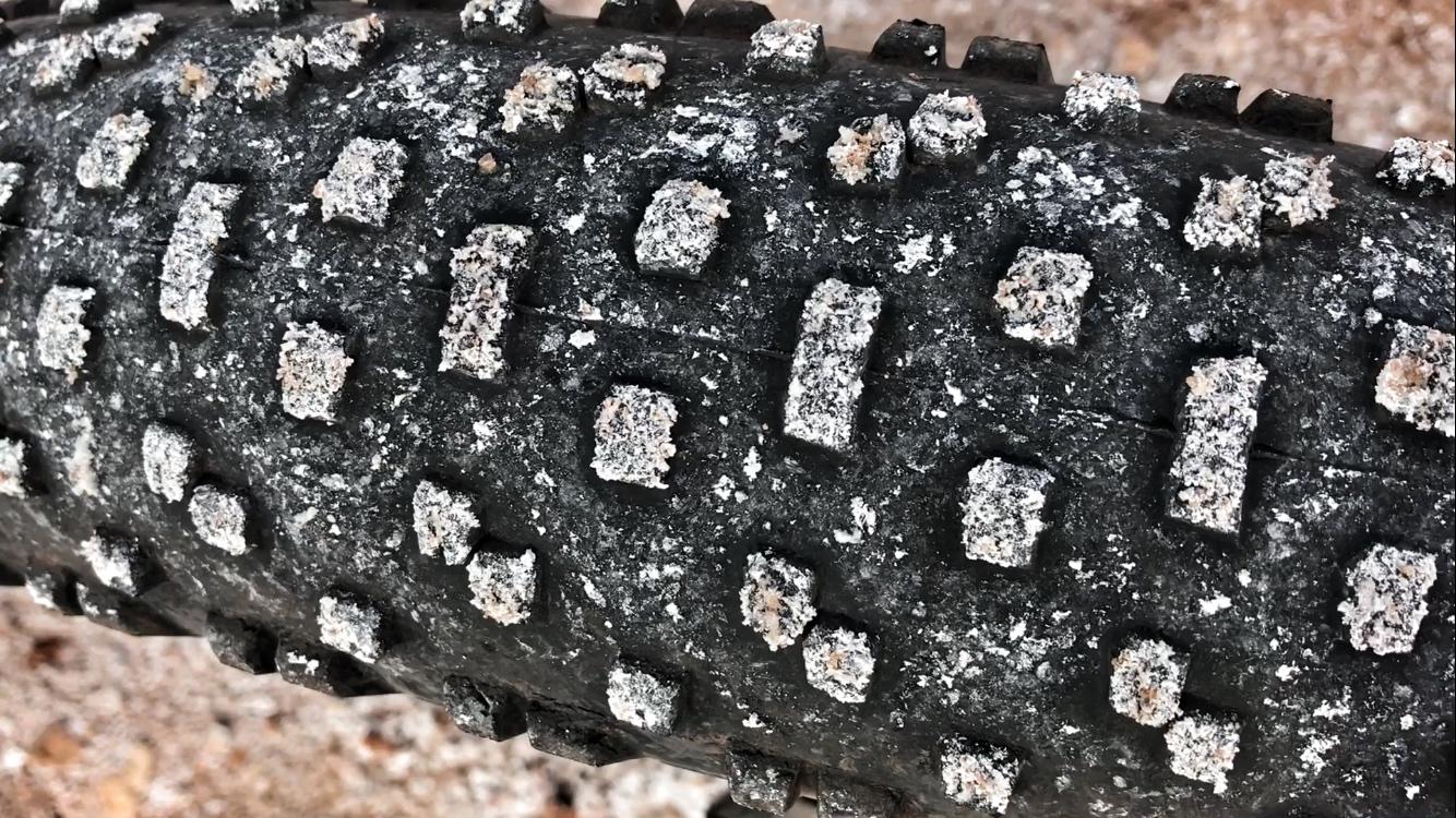 Close-up view of a textured bicycle tire tread, featuring distinct patterned knobs and speckles of salt or sand, set against a blurred, sandy background. White Ridge Bike Trails mountain bike trail.