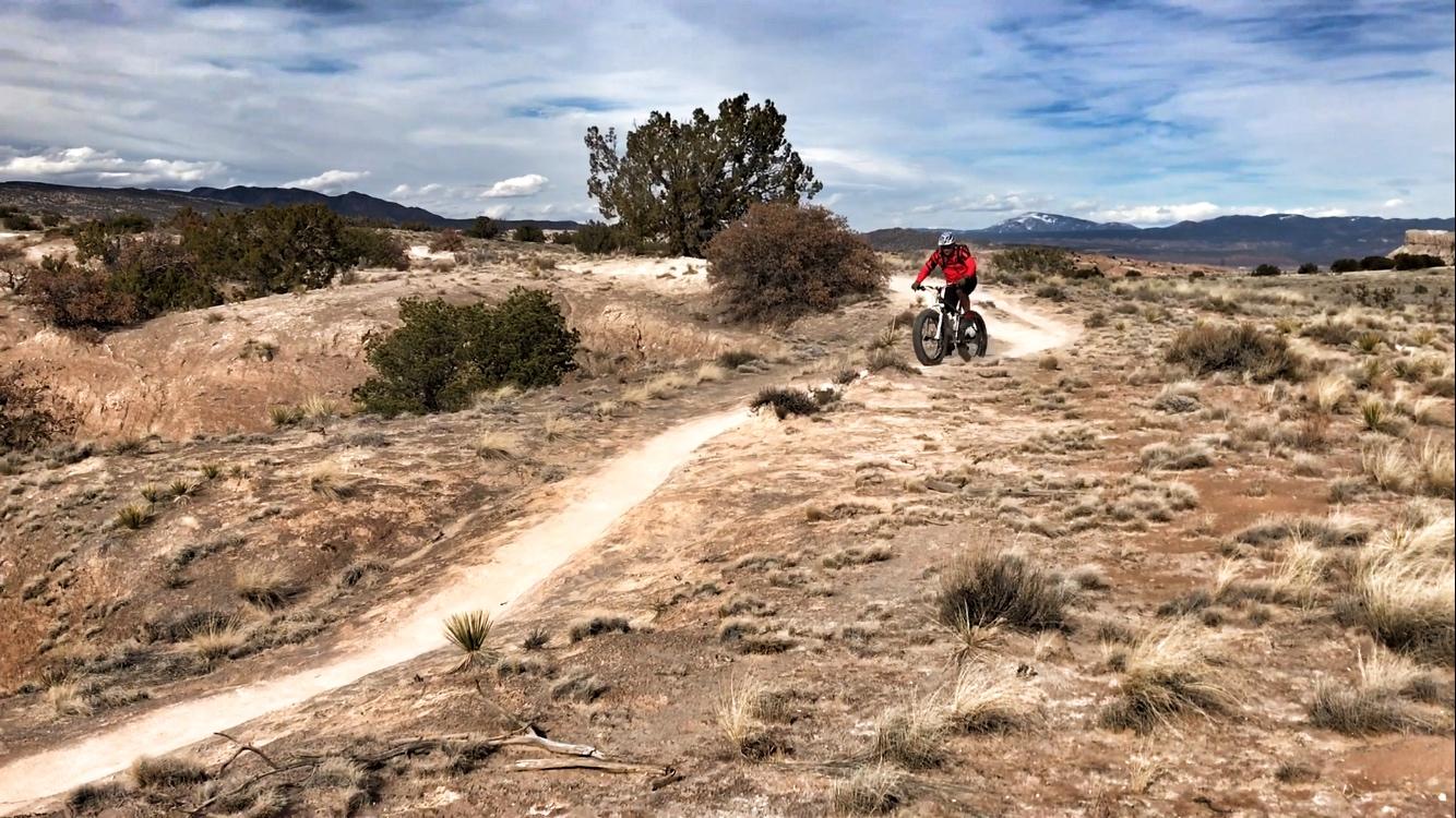 A mountain biker in a red jacket rides along a winding dirt trail through a sparse, dry landscape with low bushes and grass. In the background, mountains are visible under a partly cloudy sky. White Ridge Bike Trails mountain bike trail.