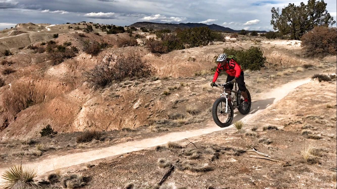 A person riding a fat bike along a dirt trail in a rugged, arid landscape. The cyclist is wearing a red jacket and a helmet, navigating through a hilly area with sparse vegetation and rocky terrain. Clouds are visible in the sky, adding to the outdoor scenery. White Ridge Bike Trails mountain bike trail.