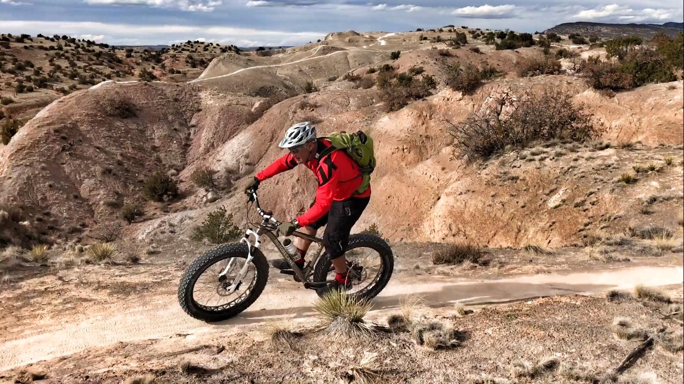 A mountain biker in a red and black outfit navigates a rocky trail in a desert landscape, with hills and sparse vegetation in the background. The biker is riding a fat bike with wide tires on a winding dirt path. White Ridge Bike Trails mountain bike trail.