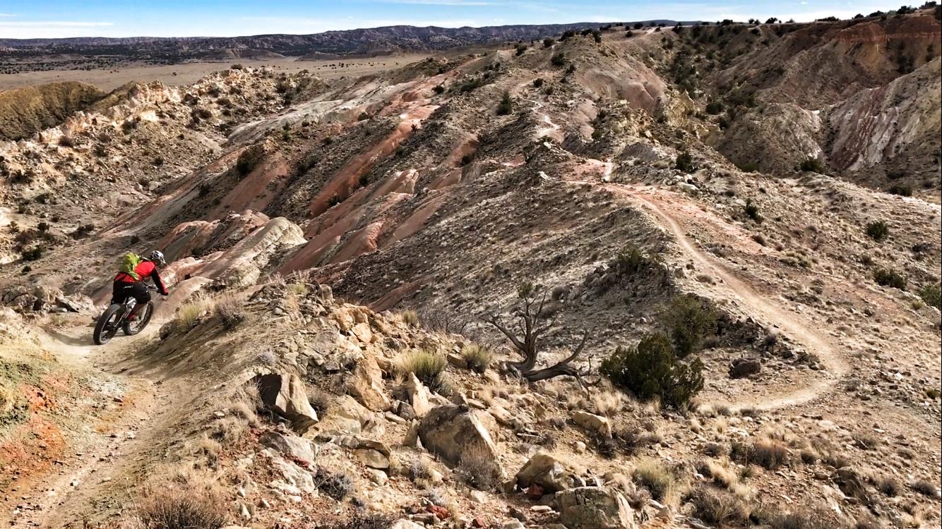 A mountain biker navigates a winding dirt trail through rugged, colorful terrain with steep slopes and sparse vegetation. The landscape features layered rock formations, shrubs, and distant hills under a clear blue sky. White Ridge Bike Trails mountain bike trail.