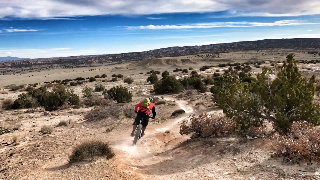 A mountain biker rides along a winding dirt trail in a rugged, desert landscape with sparse vegetation and distant hills under a partly cloudy sky. Dust is kicked up from the bike's tires as the rider descends the trail. White Ridge Bike Trails mountain bike trail.