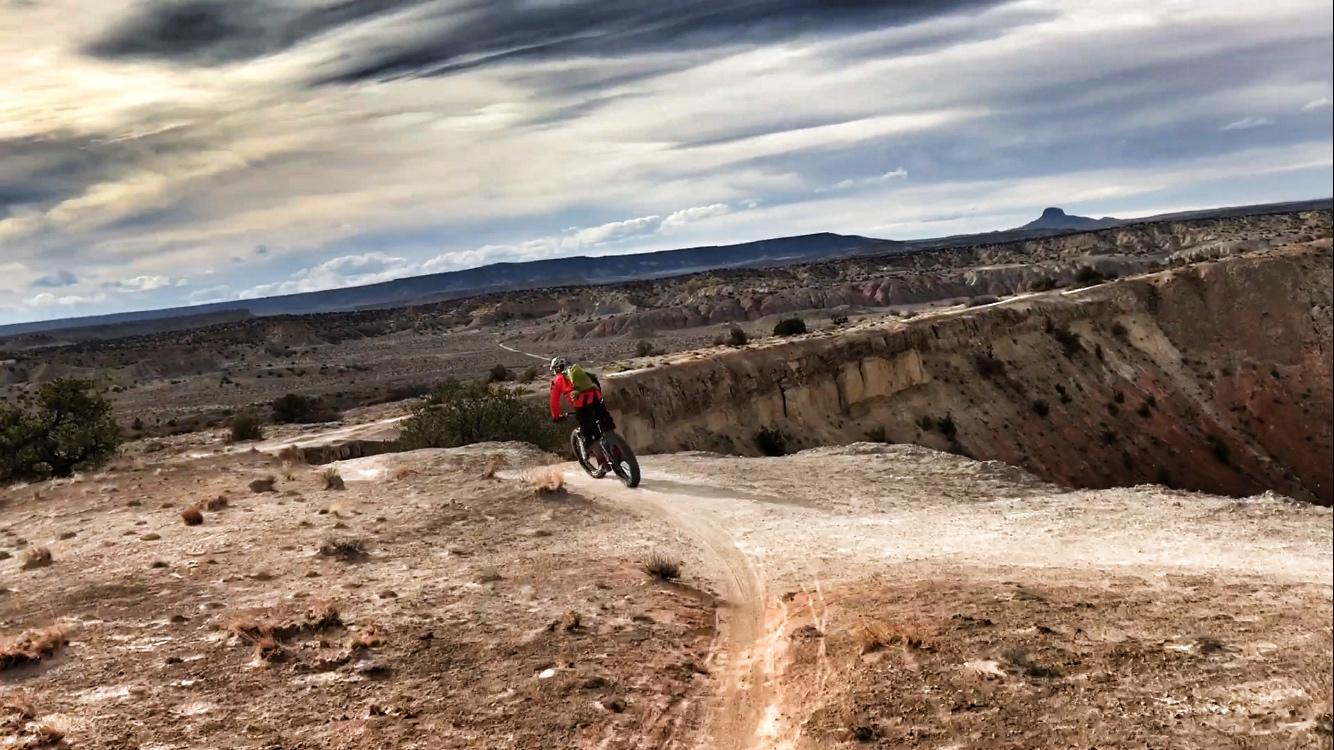 A mountain biker riding along a dirt trail in a rugged, hilly landscape under a cloudy sky. The terrain features rocky cliffs, sparse vegetation, and distant mountains on the horizon. White Ridge Bike Trails mountain bike trail.