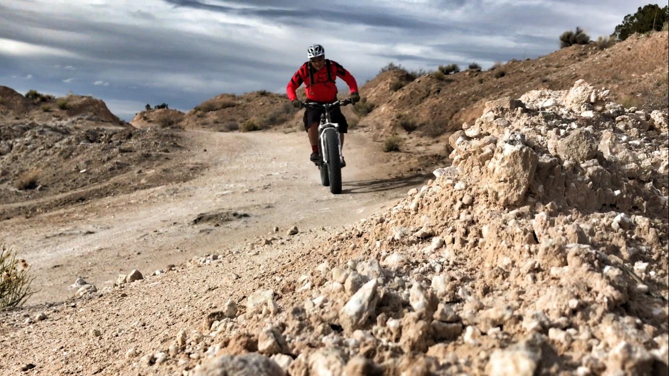 A cyclist wearing a red jacket rides a fat bike on a gravel trail surrounded by rocky terrain and sparse vegetation under a cloudy sky. The foreground features a close-up view of textured dirt and rocks. White Ridge Bike Trails mountain bike trail.