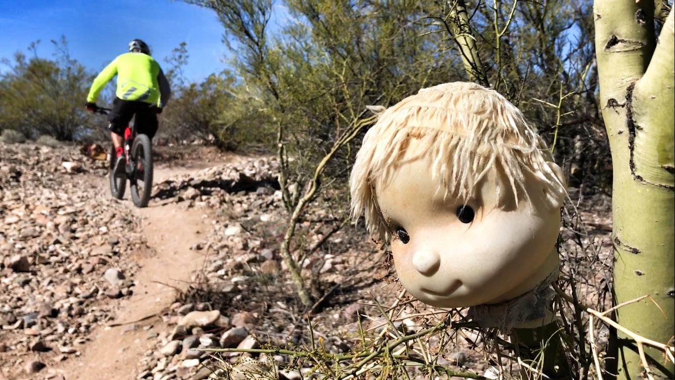 A close-up of a doll's head partially hidden among desert vegetation, with a mountain biker in the background riding along a rocky trail. The setting features sparse desert plants under a clear blue sky. Fantasy Island mountain bike trail.