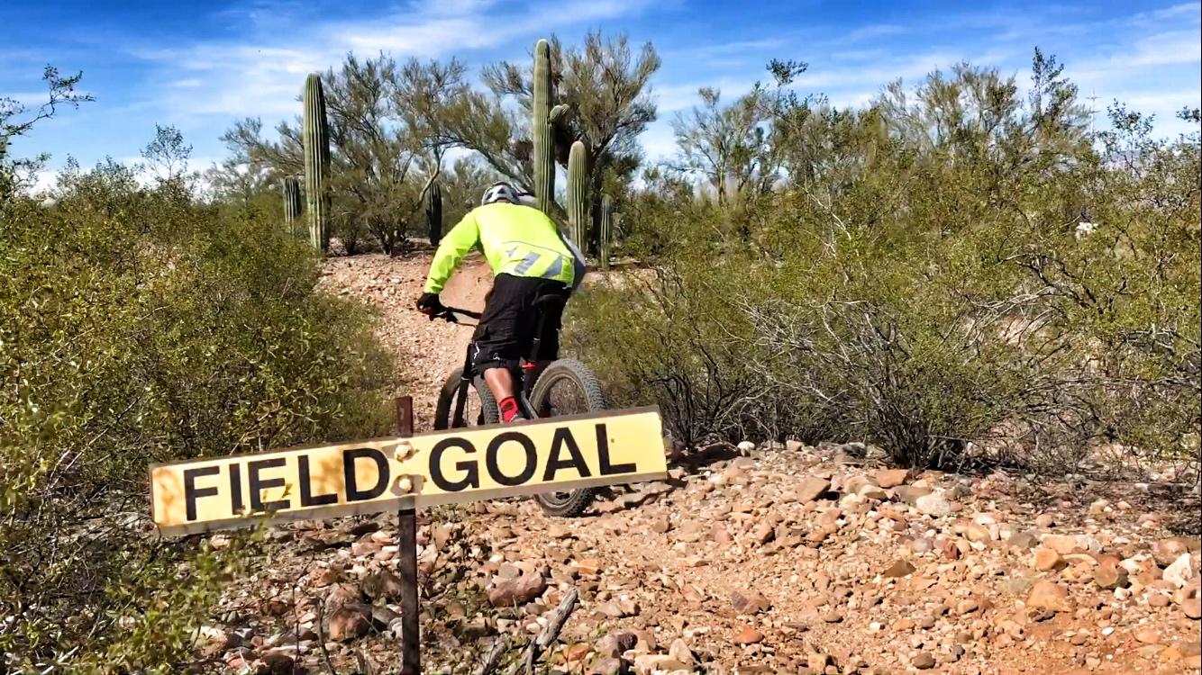 A mountain biker wearing a bright yellow jacket rides along a rocky trail in a desert landscape, passing a sign that reads "FIELD GOAL." Cacti and shrubs are visible in the background under a clear blue sky. Fantasy Island mountain bike trail.