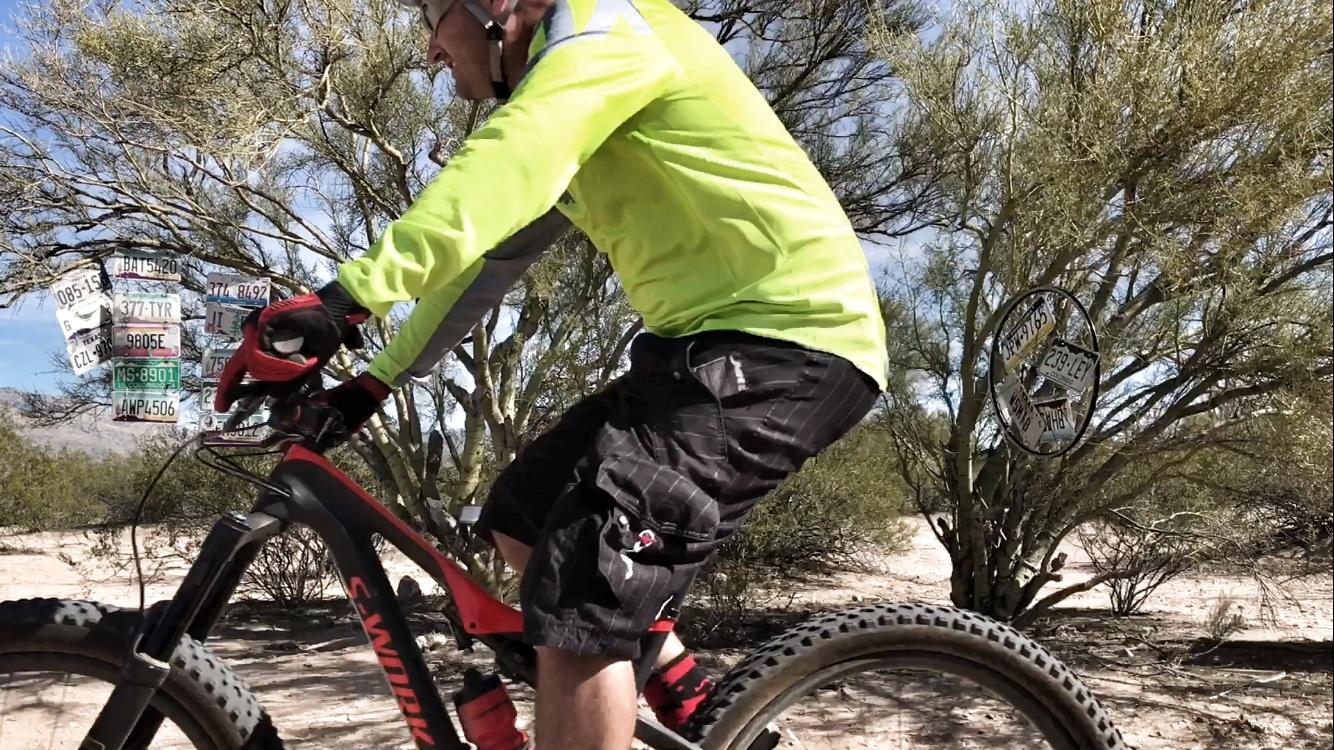 A cyclist in a bright yellow long-sleeve shirt and black shorts rides a mountain bike on a sandy trail surrounded by desert vegetation. In the background, there are several license plates affixed to a tree. The scene captures a sunny day with clear skies. Fantasy Island mountain bike trail.