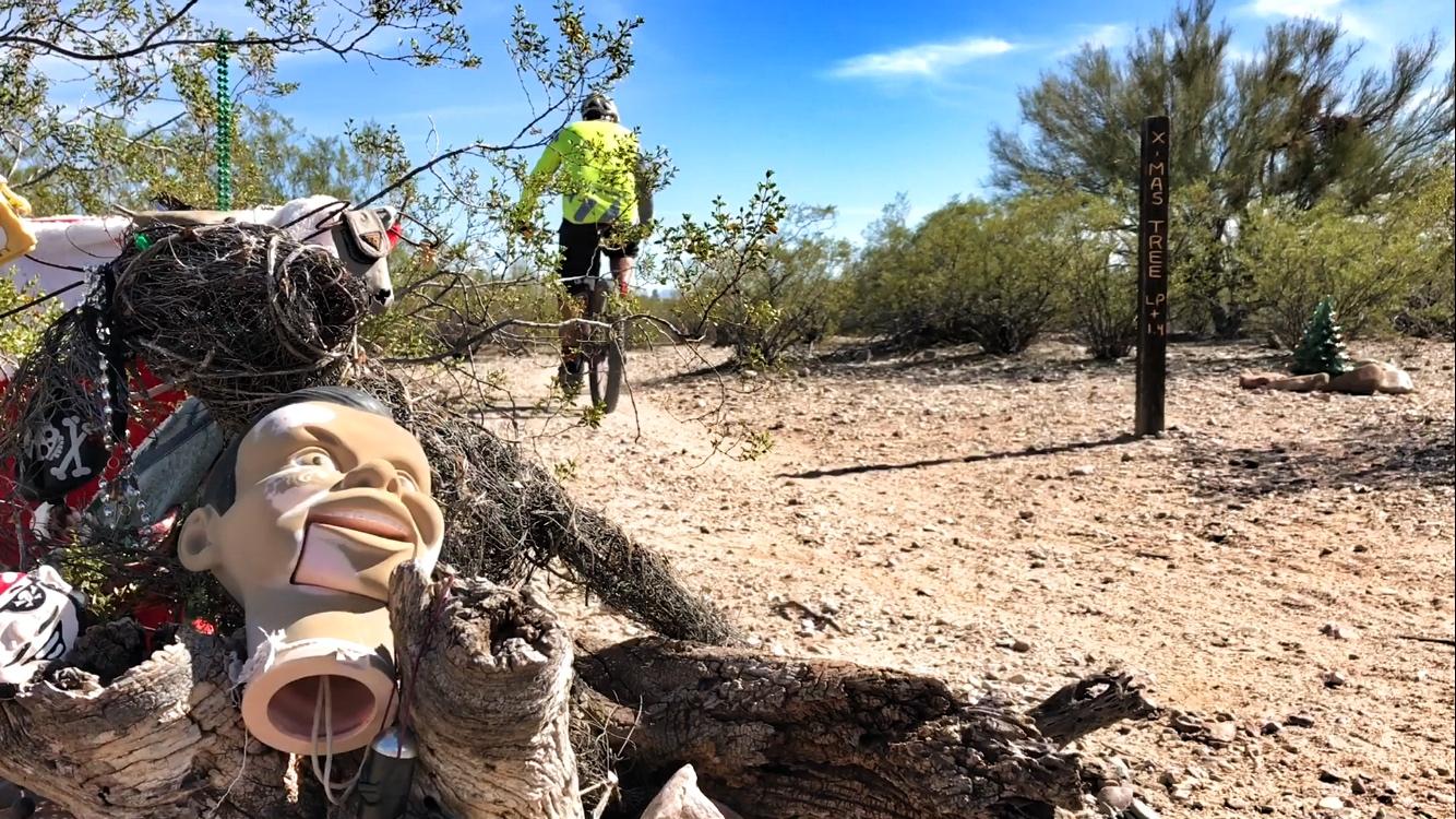 A desert scene featuring a humorous sculpture of a cartoonish head mounted on a piece of wood, surrounded by various decorations like beads and pirate-themed items. In the background, a cyclist in bright clothing rides along a dirt path, with a trail marker labeled "X-MAS TREE" visible nearby. The landscape is dotted with sparse bushes and clear blue skies. Fantasy Island mountain bike trail.