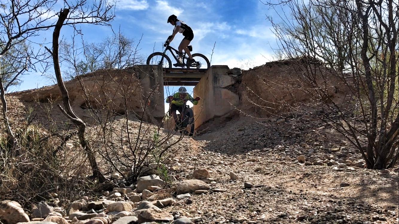 A mountain biker navigating a rocky trail while another rider is seen biking over a concrete bridge in the background, surrounded by sparse vegetation and a clear blue sky. Fantasy Island mountain bike trail.