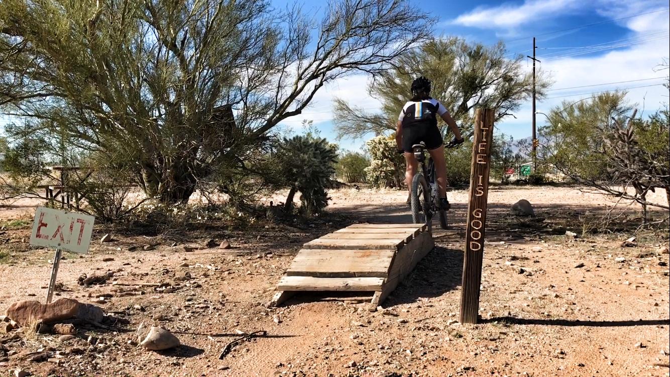 A mountain biker riding over a wooden ramp on a desert trail, with a sign next to them that reads "LIFE IS GOOD" and another sign in the background indicating "EXIT." The scene features sparse desert vegetation and a clear blue sky. Fantasy Island mountain bike trail.