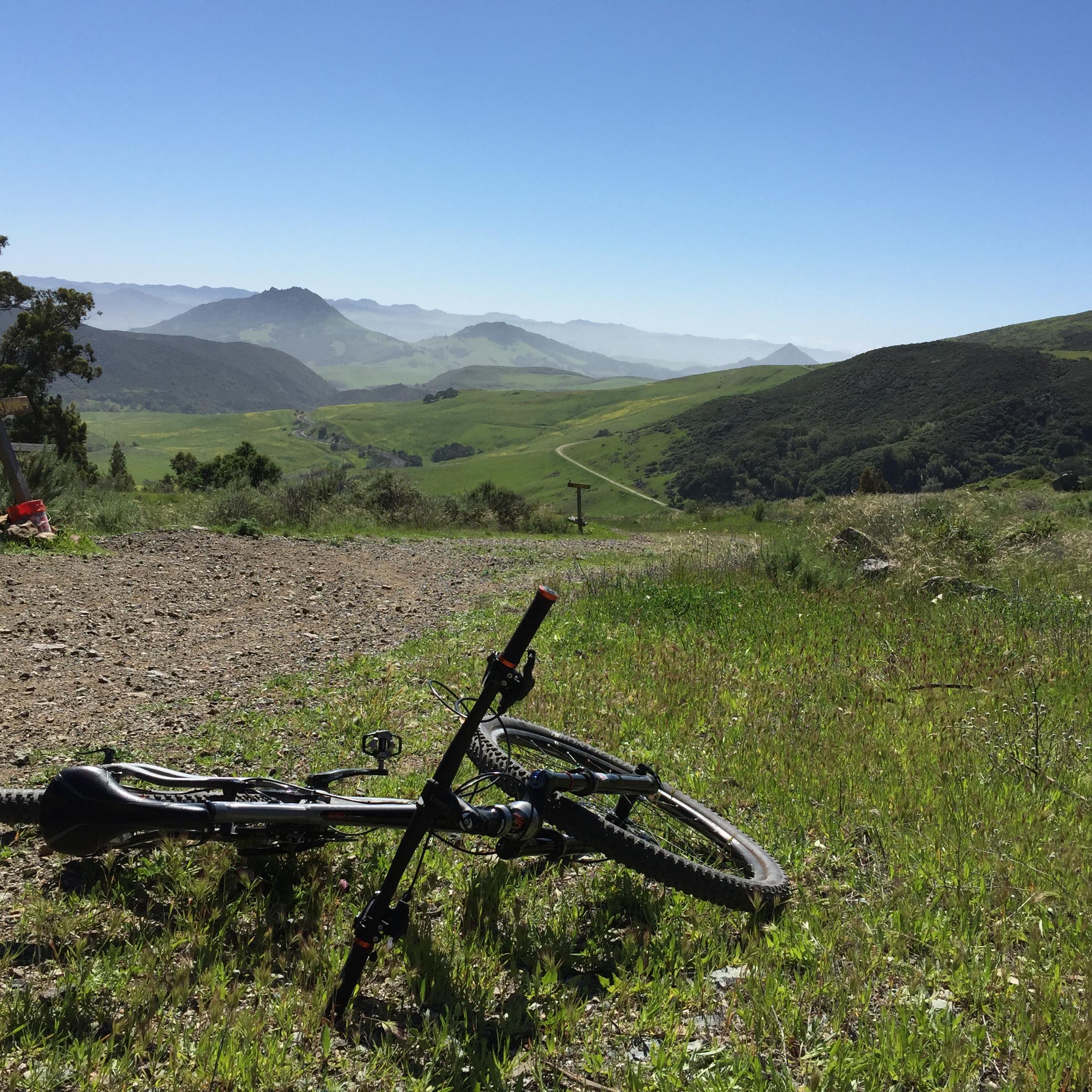Trek Remedy 9: A mountain bike rests on a gravel path overlooking a lush green valley and rolling hills under a clear blue sky. In the background, misty mountains rise in the distance, creating a serene outdoor landscape.
