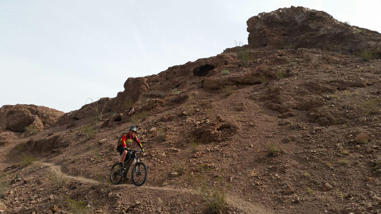 A mountain biker navigating a rocky trail on a hillside, surrounded by dry terrain and sparse vegetation under a clear sky. Sugarloaf Peak mountain bike trail.