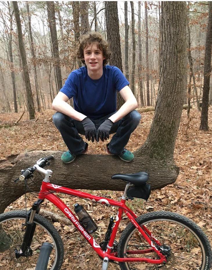 Gary Fisher Piranha: A young person with curly hair is sitting on a fallen tree in a forest, wearing a blue shirt and black gloves. They are smiling and posed casually with their legs extended. A red mountain bike is leaning against the tree nearby, surrounded by dry leaves and trees in the background.