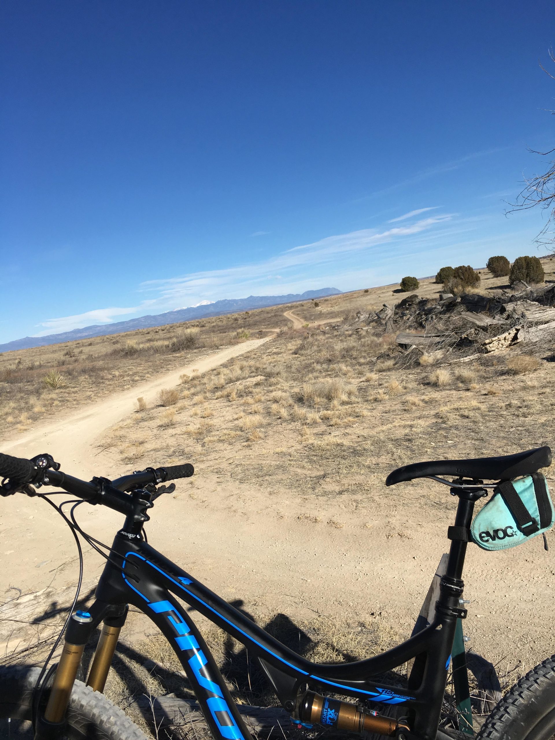 A mountain bike rests on a rocky outcrop, overlooking a winding dirt path that leads into a vast, open landscape. The scene features a clear blue sky with a few wispy clouds and distant mountains in the background. Sparse vegetation and dry grass cover the foreground, suggesting a rugged outdoor environment. South Shore Lake Pueblo mountain bike trail.