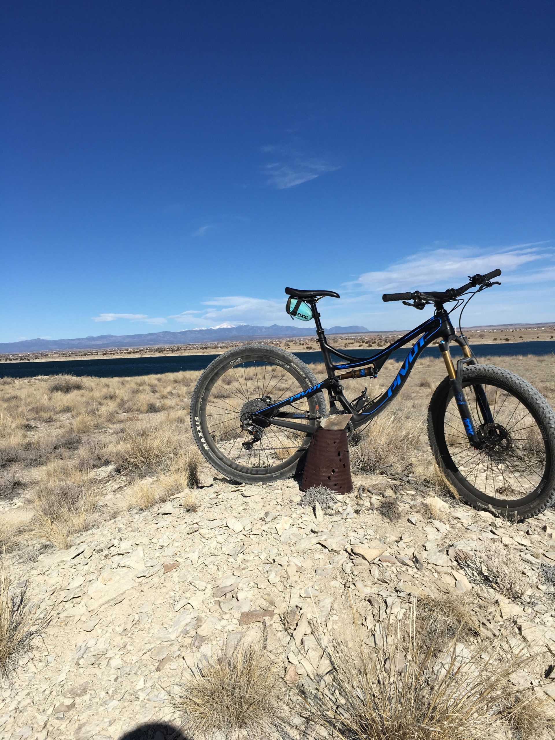 A mountain bike rests on a rocky outcrop in a barren landscape, with a clear blue sky and distant mountains in the background. The foreground features dry grasses and a small, rusted object partially buried in the ground. South Shore Lake Pueblo mountain bike trail.